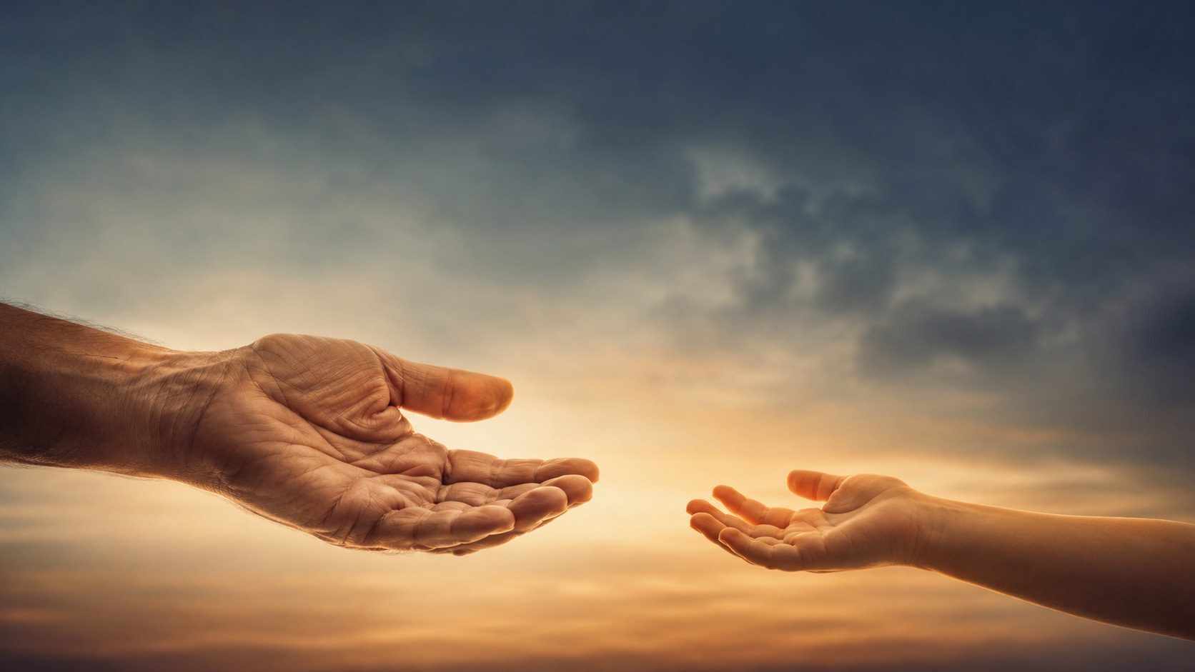 An adult hand and a child's hand reaching toward each other at sunset.